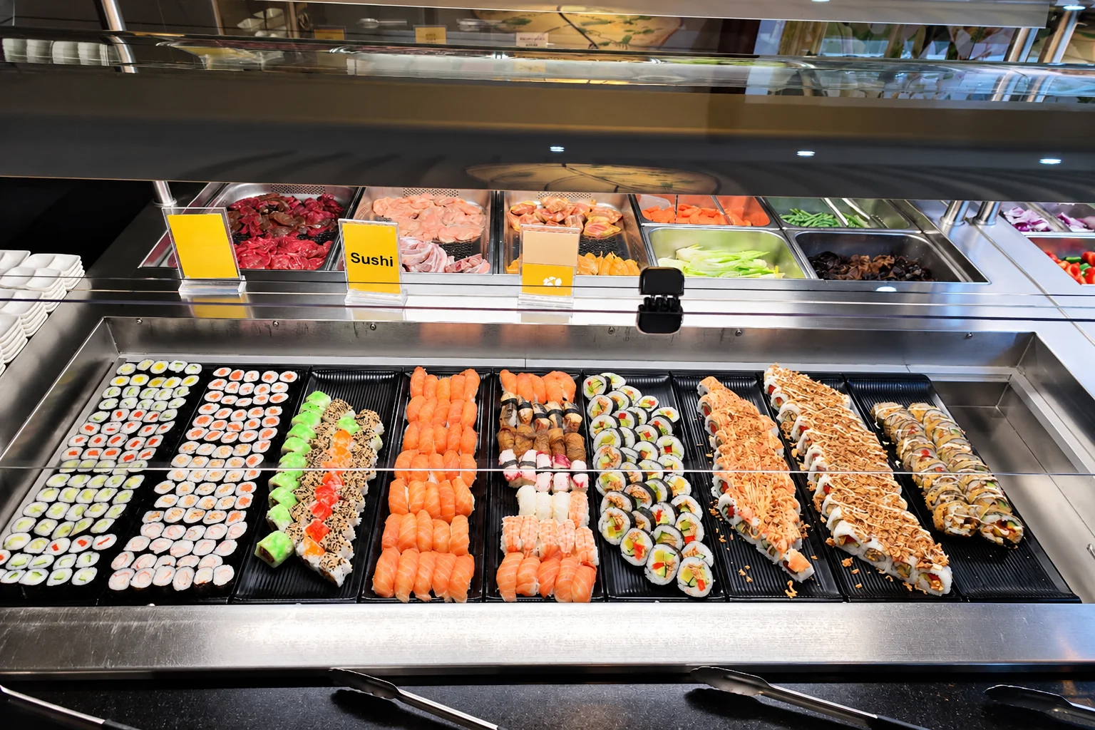 Sushi-Theke am Büffet: Reihen von Maki, Inside-Out-Rollen und Nigiri mit Lachs auf schwarzen Tabletts, ein gelbes Schild „Sushi“.