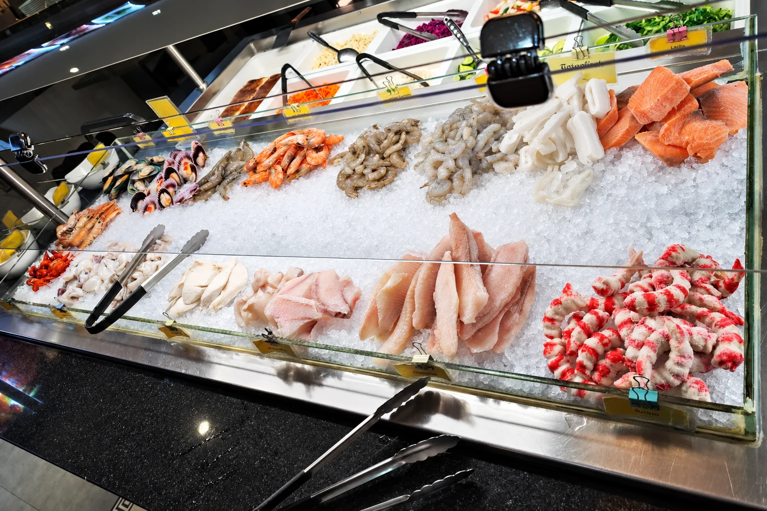 Meeresfrüchte auf Eis am Büffet: Garnelen, Lachs, Tintenfisch, Fischfilets und Surimi, davor Zangen.