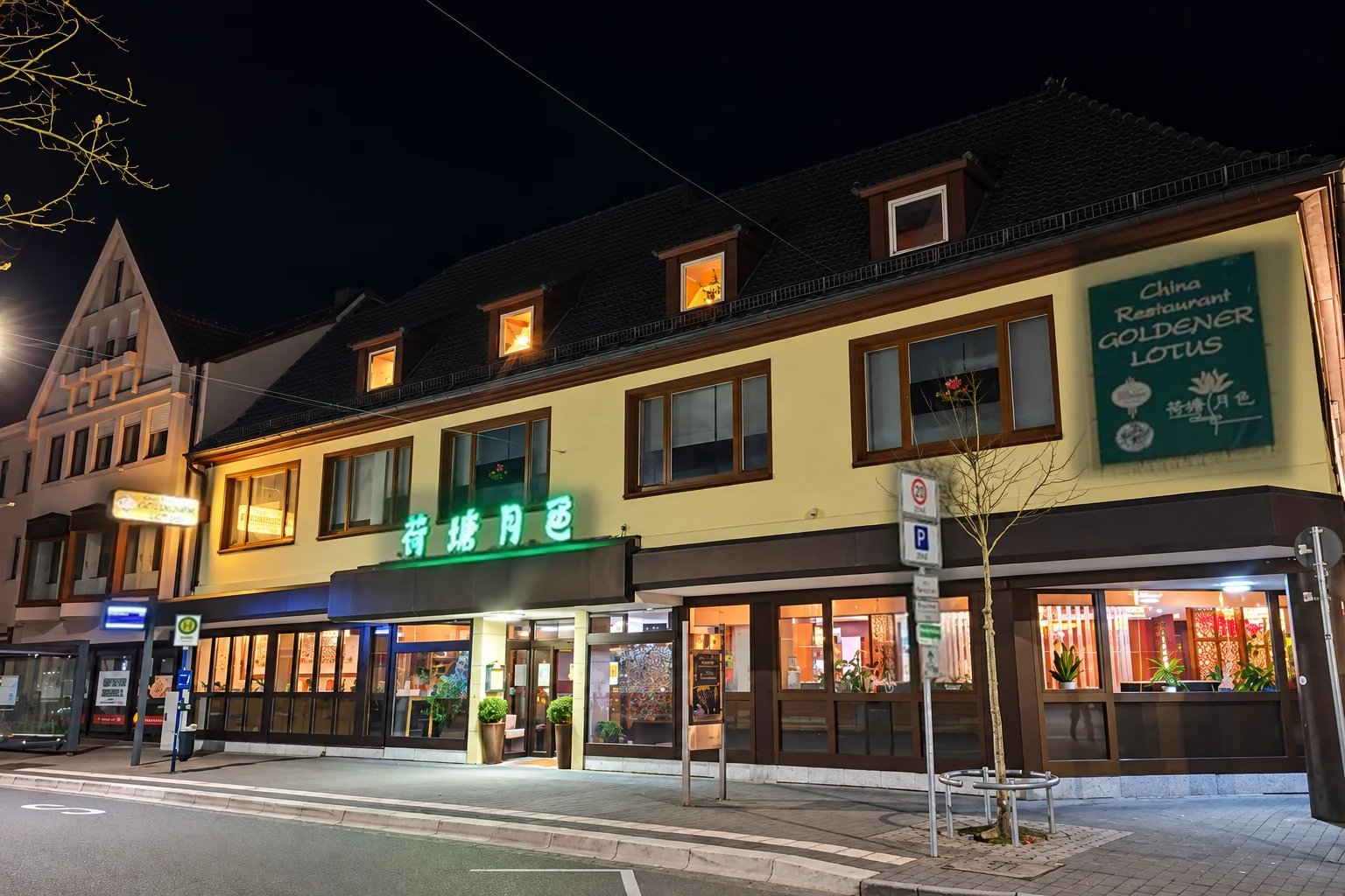 Die Fassade des Goldener Lotus am Abend: gelbes Haus, grüne Leuchtschrift über dem Eingang, Schild „China Restaurant Goldener Lotus“, beleuchtete Fenster.